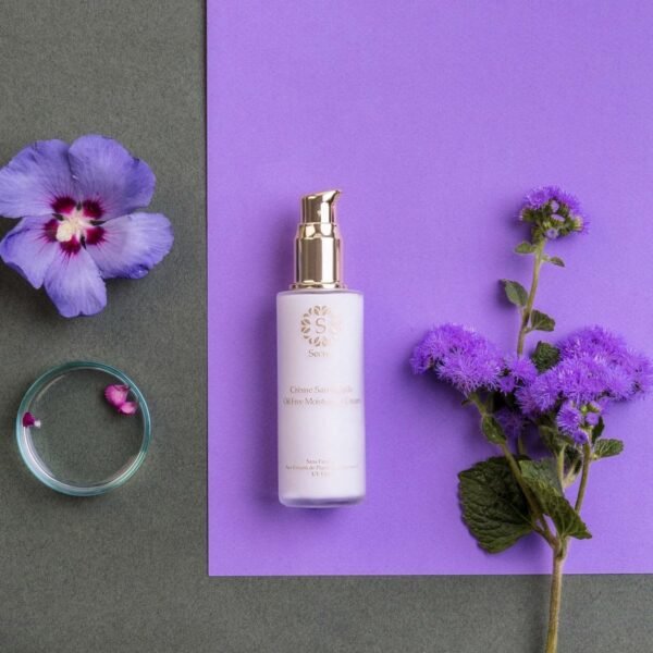 White cosmetic bottle with a gold pump sits on a purple surface, flanked by purple flowers and a small glass dish with petals nearby.