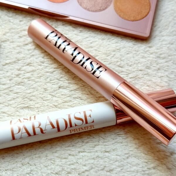 Rose-gold mascara tube labeled 'PARADISE' next to a pink makeup palette on a soft textured surface, suggesting a beauty routine.