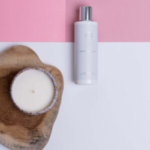 Pale skincare bottle with a silver cap on a pink-and-white backdrop beside a wooden slab with a round candle in a decorative tin.