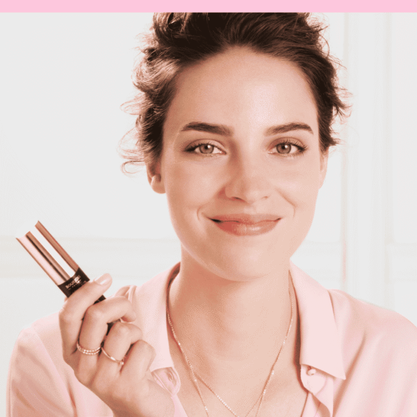  BEAUTÉNIUM Close up of a smiling woman holding a pink lipstick near her lips wearing a pink blouse