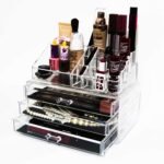 Clear acrylic makeup organizer filled with lipsticks, nail polishes, brushes and jewelry in a multi-drawer unit on a white background.